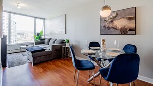 A modern living room with a sofa, dining table, chairs, and a city view. - 1BR Condo Near Rogers Arena | Heart of Vancouver (Vancouver)