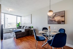 A modern living room with a sofa, dining table, chairs, and a city view. - 1BR Condo Near Rogers Arena | Heart of Vancouver (Vancouver)