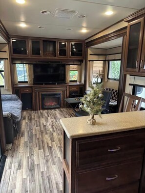 A well-lit RV interior with a TV, fireplace, dining area, and a small Christmas tree.