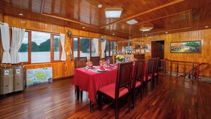 Interior de madera con una mesa de comedor dispuesta para seis personas, ventanas amplias con vistas a un cuerpo de agua y una zona de bar con botellas.