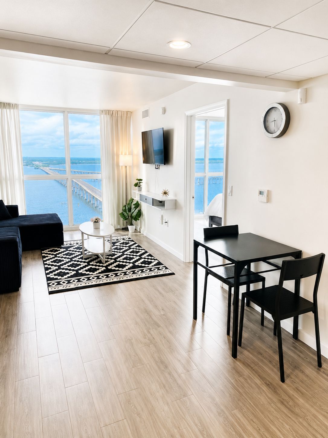 A modern living room with a large window, a flat-screen TV, a dining area with a table and chairs, and a view of a bridge and water.