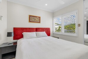 Un dormitorio con cama, mesita de noche, lámpara, cuadro en la pared y ventana con persianas.