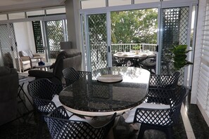 Ein sonnendurchfluteter Patio-Bereich mit einem runden Tisch und Stühlen, eine überdachte Sitzgelegenheit und ein Blick ins Freie durch eine Glasdoor.