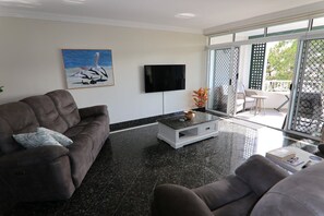Ein modernes Wohnzimmer mit einer Couch, einem Couchtisch und einem Fernseher. Ein Balkon bietet Blick ins Freie.