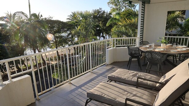 Een overdekt terras met loungezetels en een tafel, omgeven door een witte balustrade, met uitzicht op een aangelegde tuin met palmboompjes.
