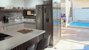 Une cuisine moderne dotée d’un îlot central, d’un coin repas avec des chaises, d’un réfrigérateur et d’une porte vitrée donnant sur l’espace piscine.