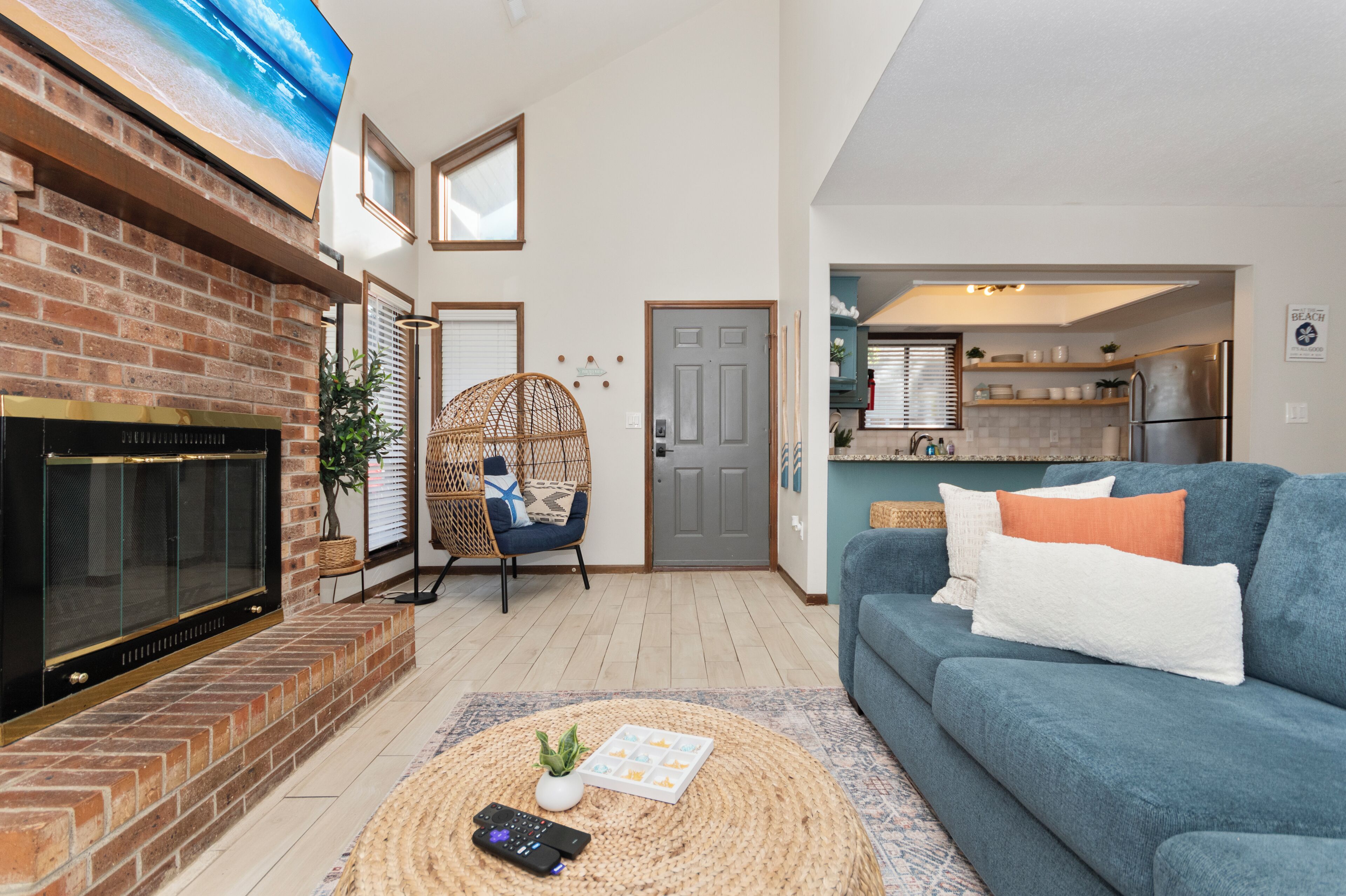 A living room with a fireplace, a blue sofa, a wicker chair, and a kitchen visible through an open door.