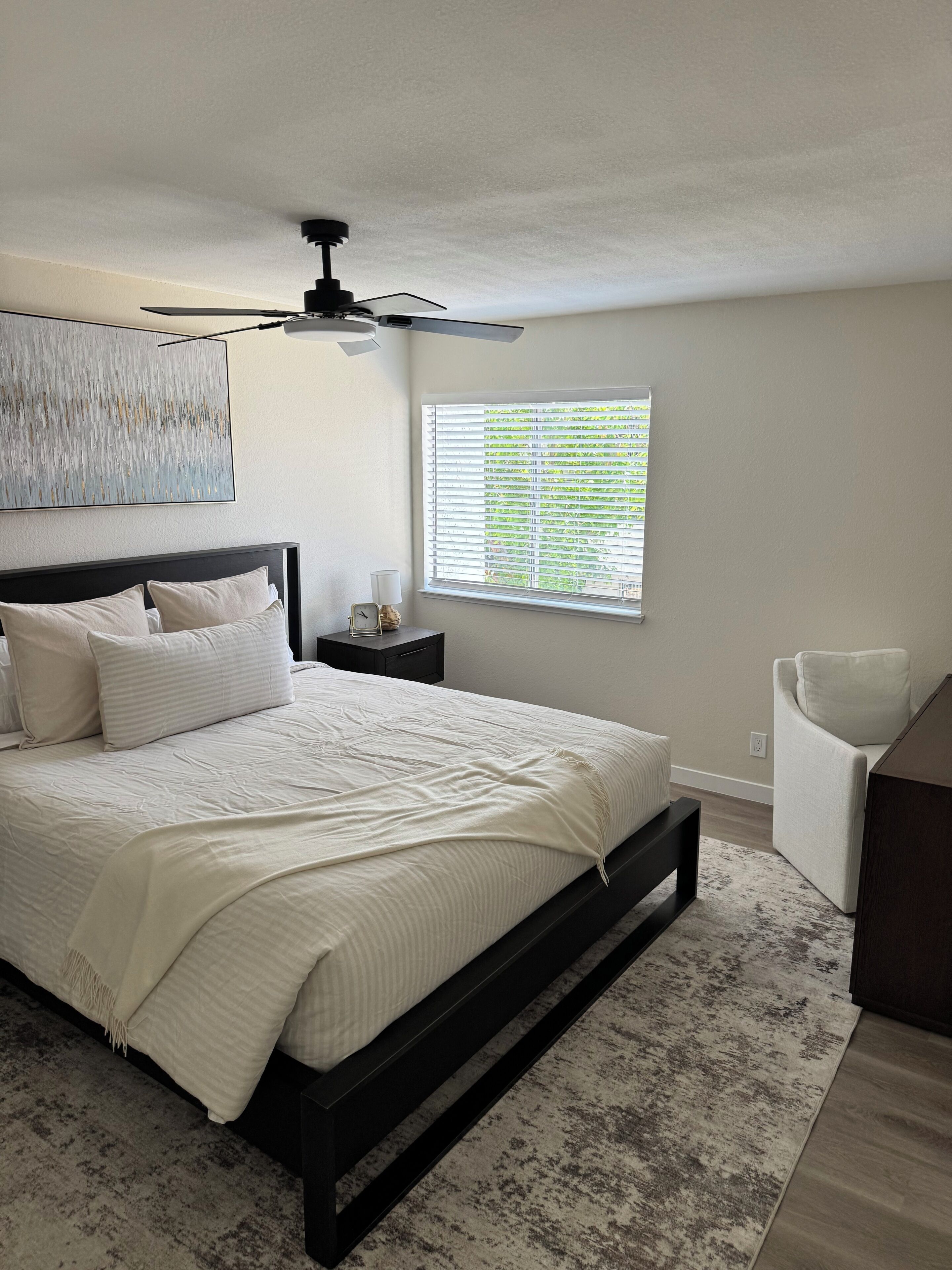 A bedroom with a bed, a nightstand, a ceiling fan, a window with blinds, and a chair.