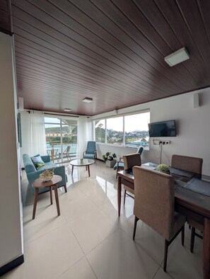 En moderne stue med træloft, et fladskærms-tv monteret på væggen, en sofa, et spisebord med stole og en balkon med udsigt over byen.