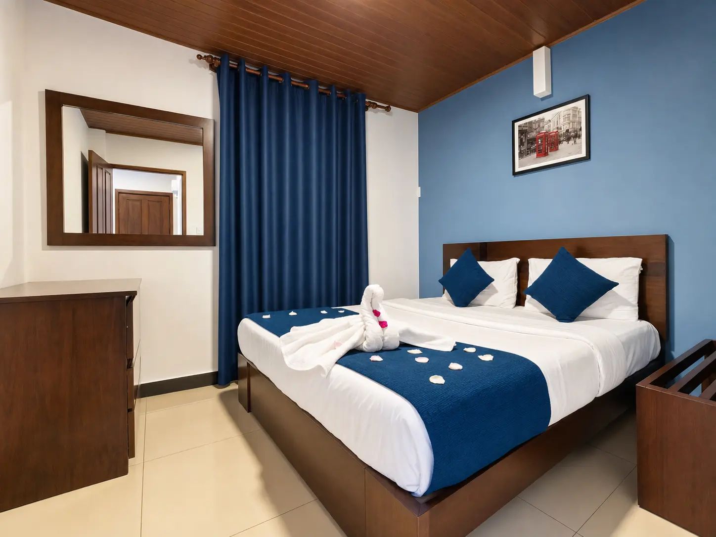 Habitación de hotel con cama, cabecera de madera, cortina azul, un cuadro en la pared y cómoda de madera.
