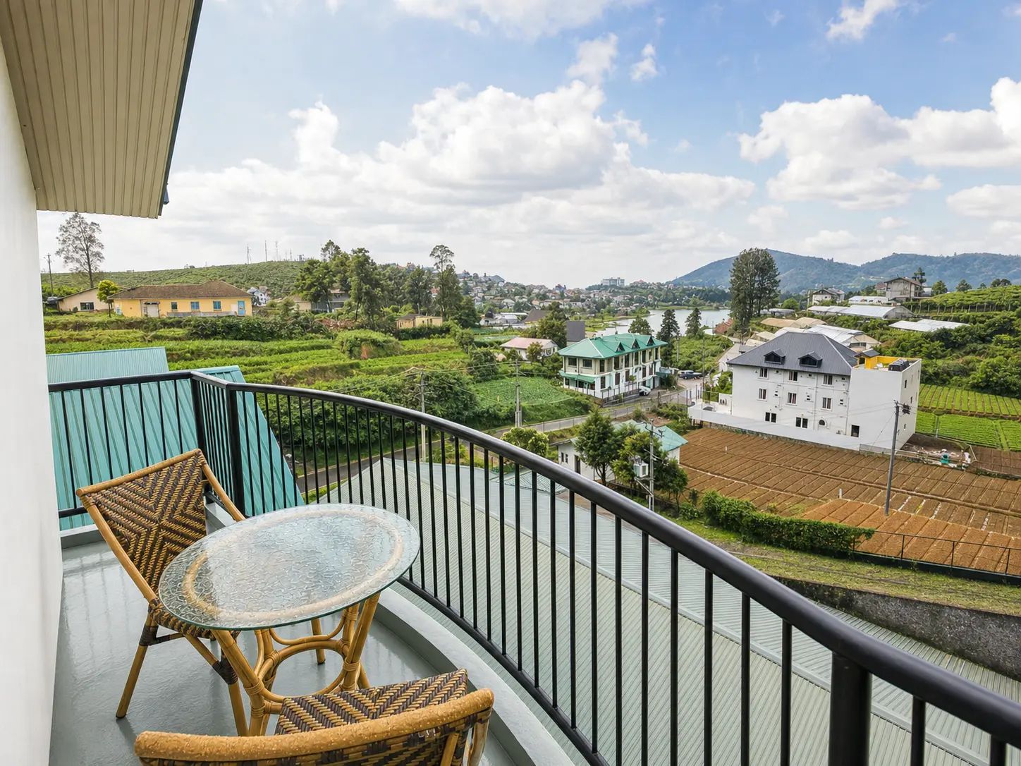 A balcony with wicker furniture and a glass table, overlooking a residential area with houses and green fields.