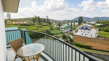 A balcony with wicker furniture and a glass table, overlooking a residential area with houses and green fields.