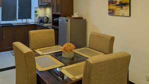 Un comedor con mesa de madera, cuatro sillas, un jarrón con flores, una cocina al fondo y un cuadro en la pared.