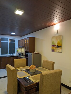 A dining area with a wooden table, four chairs, a vase with flowers, a kitchen in the background, and a painting on the wall.