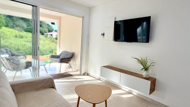 A modern living room with a sofa, a small wooden table, a TV mounted on the wall, and a sliding glass door leading to an outdoor area. - The cos cocoon, less than 10 minutes from the airport. (Ducos)