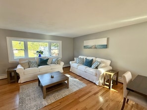 A living room with two white sofas, a coffee table, a dining table with chairs, a small side table, and a painting on the wall. - Coastal Bristol, RI 4-Bedroom Home Near Downtown, RWU, & Wedding Venues (Bristol)