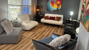 Ein Wohnzimmer mit einem weißen Sofa, einem grauen Sessel und einem schwarzen Stuhl. Es gibt einen Holzboden, einen Deckenventilator und ein Gemälde an der Wand.