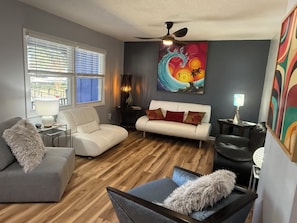 A living room with a white sofa, grey armchair, and a black chair. There is a wooden floor, a ceiling fan, and a painting on the wall. - Sunny & SAndy Surf Social , 3/2 Mid Century Vibes ,Sunset Vibes, Coastal Stays (Myrtle Beach)