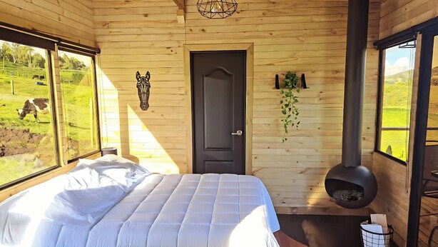 A wooden cabin bedroom with a bed, a door, a hanging plant, a fireplace, and a window with a view of cows grazing. - Cabaña chalet  con chimenea con wi-fi para nómadas digitales (Medellín)