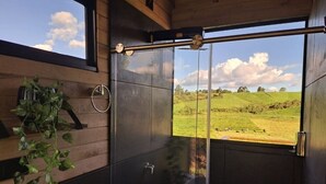 A modern bathroom with a glass shower enclosure, a toilet, and a large window offering a view of a green landscape. - Cabaña chalet  con chimenea con wi-fi para nómadas digitales (Medellín)