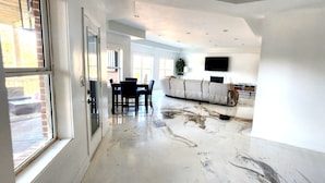 A spacious living area with a large window, a dining table with chairs, a sofa, and a flat-screen TV mounted on the wall.