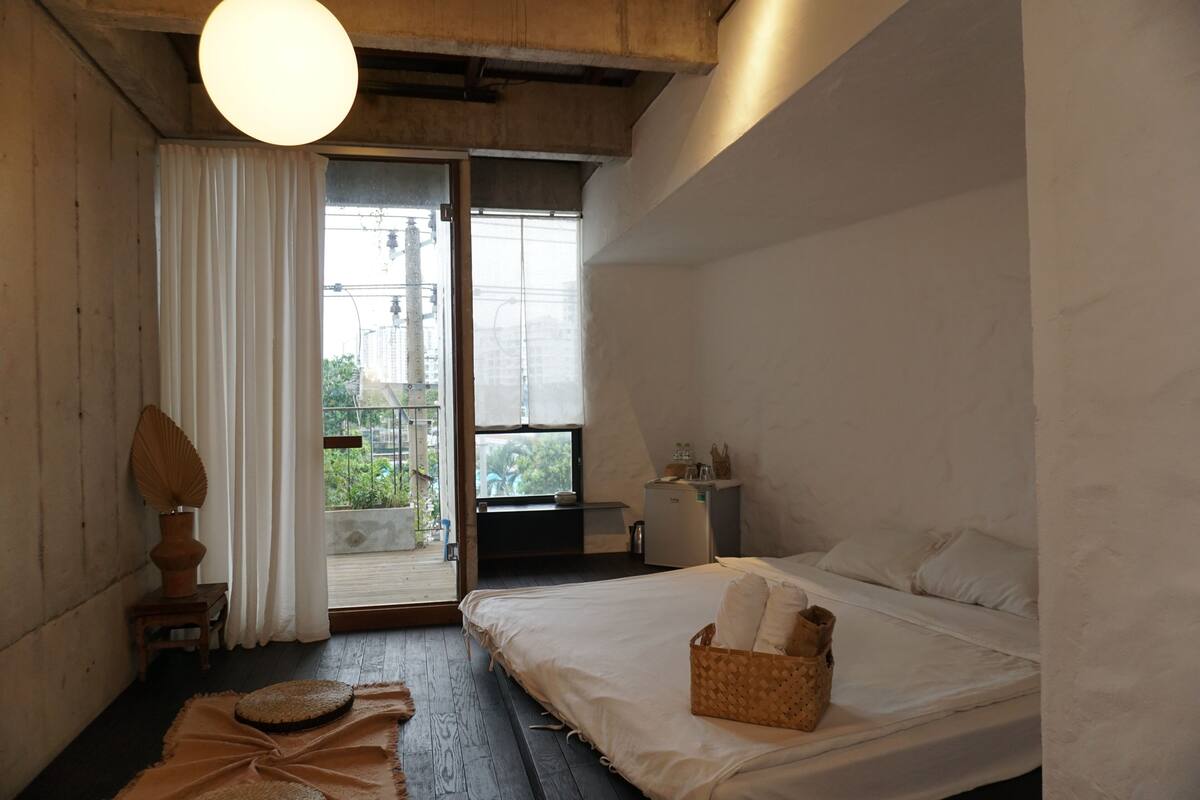 A bedroom with a bed, a small table, a lamp, a window, and a balcony.