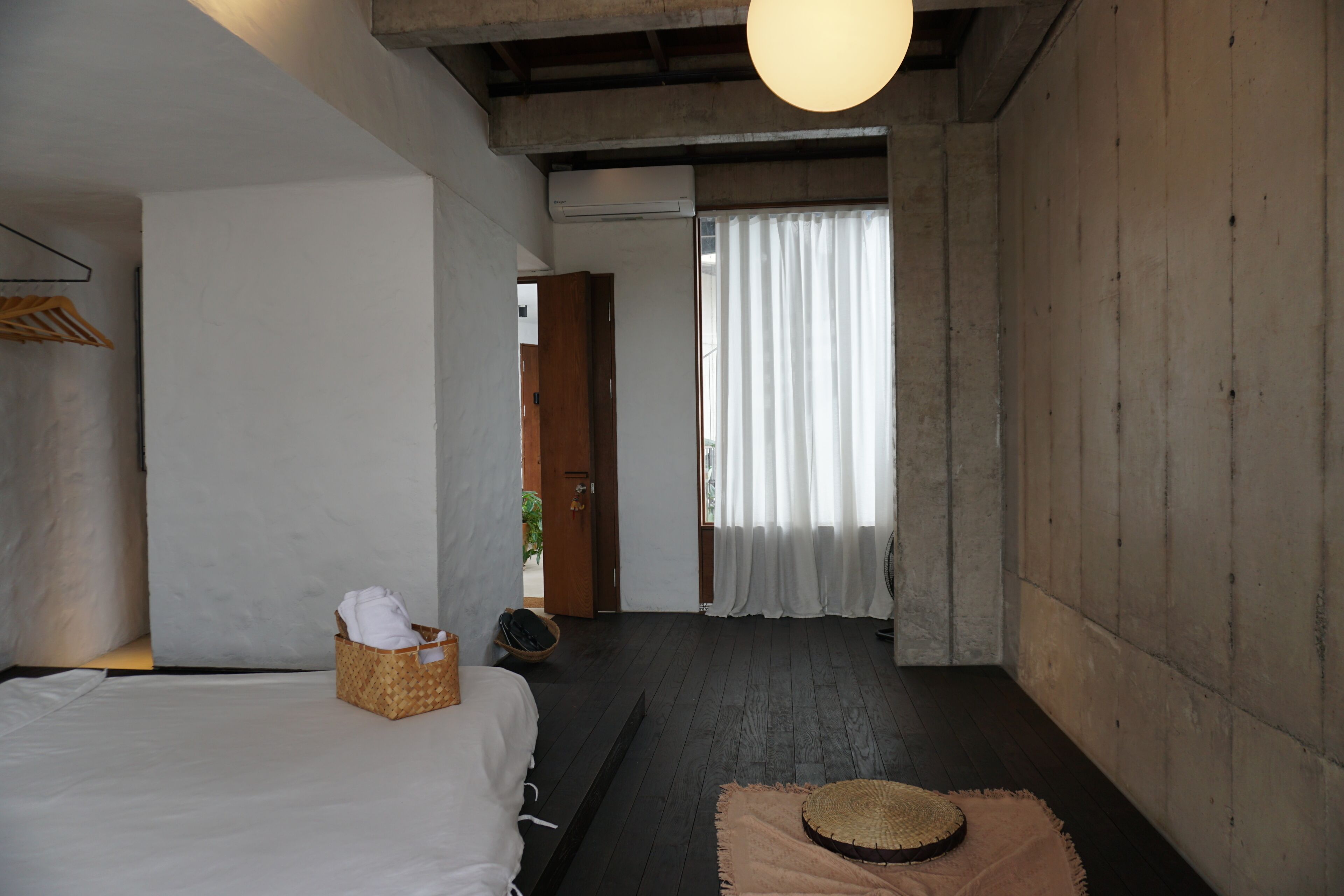 Une chambre avec un lit, un panier en osier, un ventilateur et une vue sur l’extérieur, visible à travers une fenêtre ornée de rideaux.
