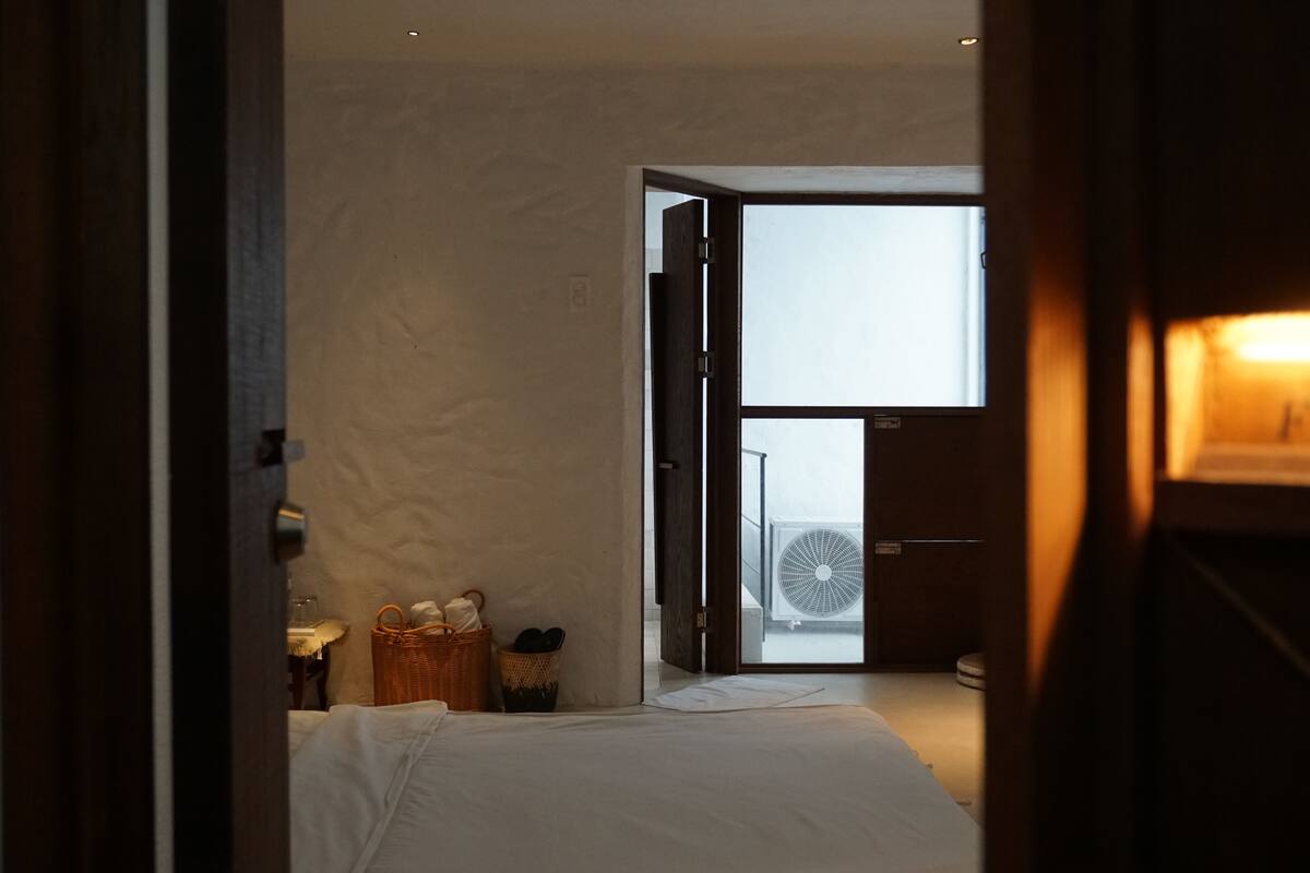 A bedroom with a bed, a wicker basket, a nightstand, and a window with blinds.