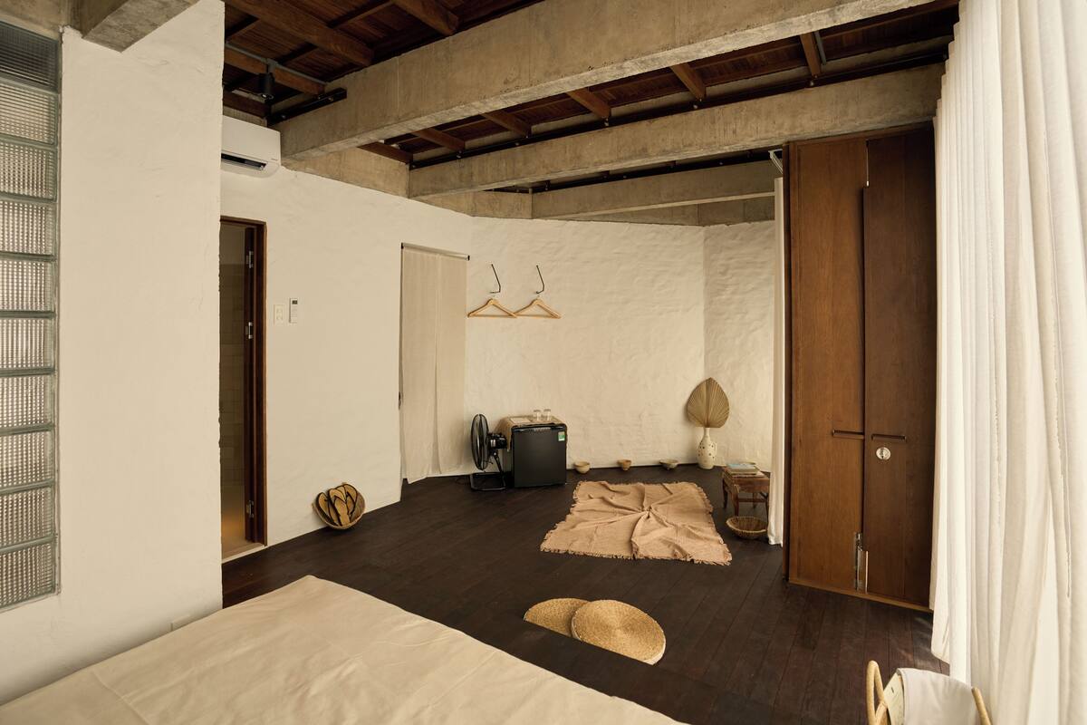 A room with a bed, a wooden wardrobe, a fan, and a small table with a lamp.