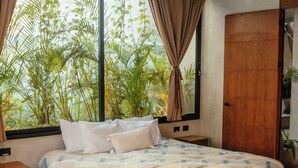A bedroom with a large bed, a ceiling fan, a window with curtains, and a view of greenery outside.