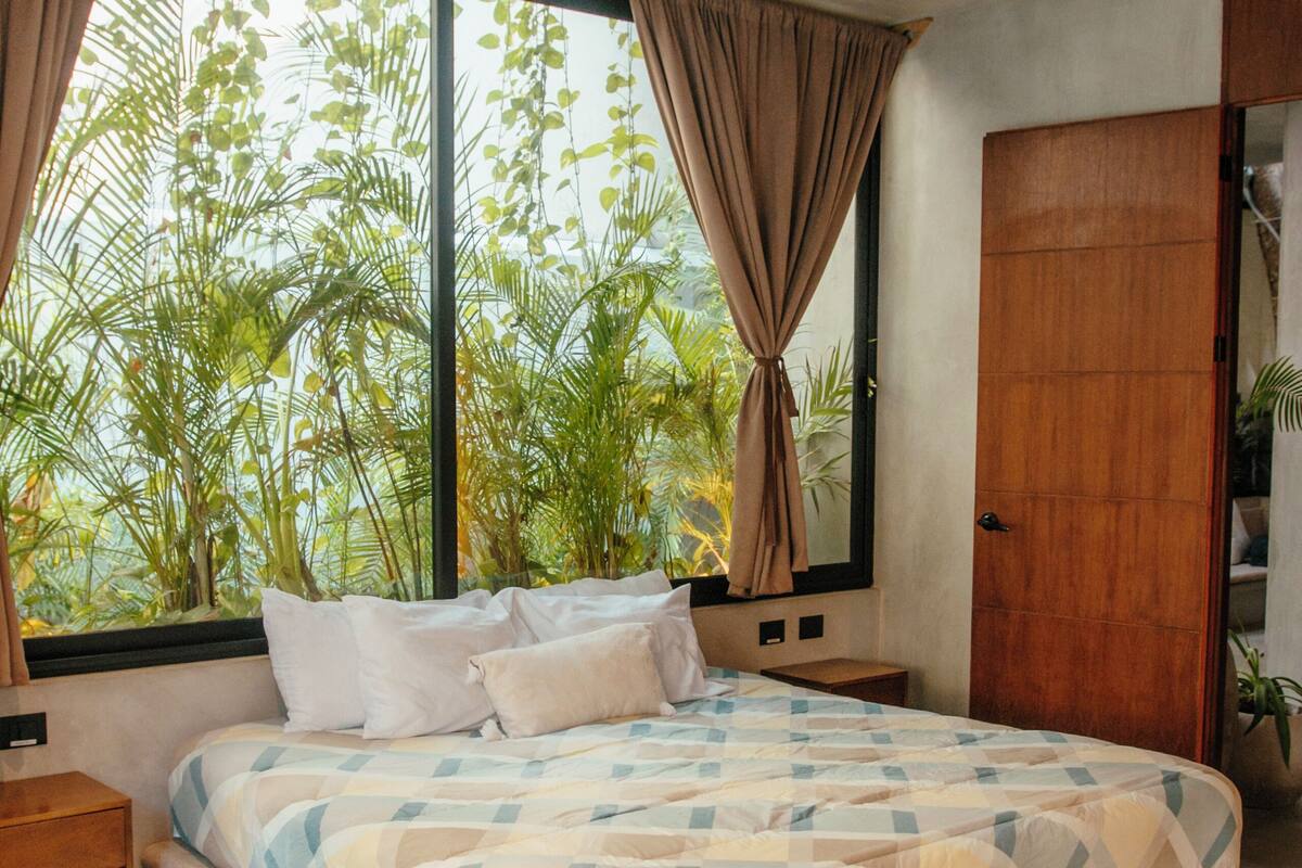 Un dormitorio con una cama grande, un ventilador de techo, una ventana con cortinas y vistas al exterior con vegetación.