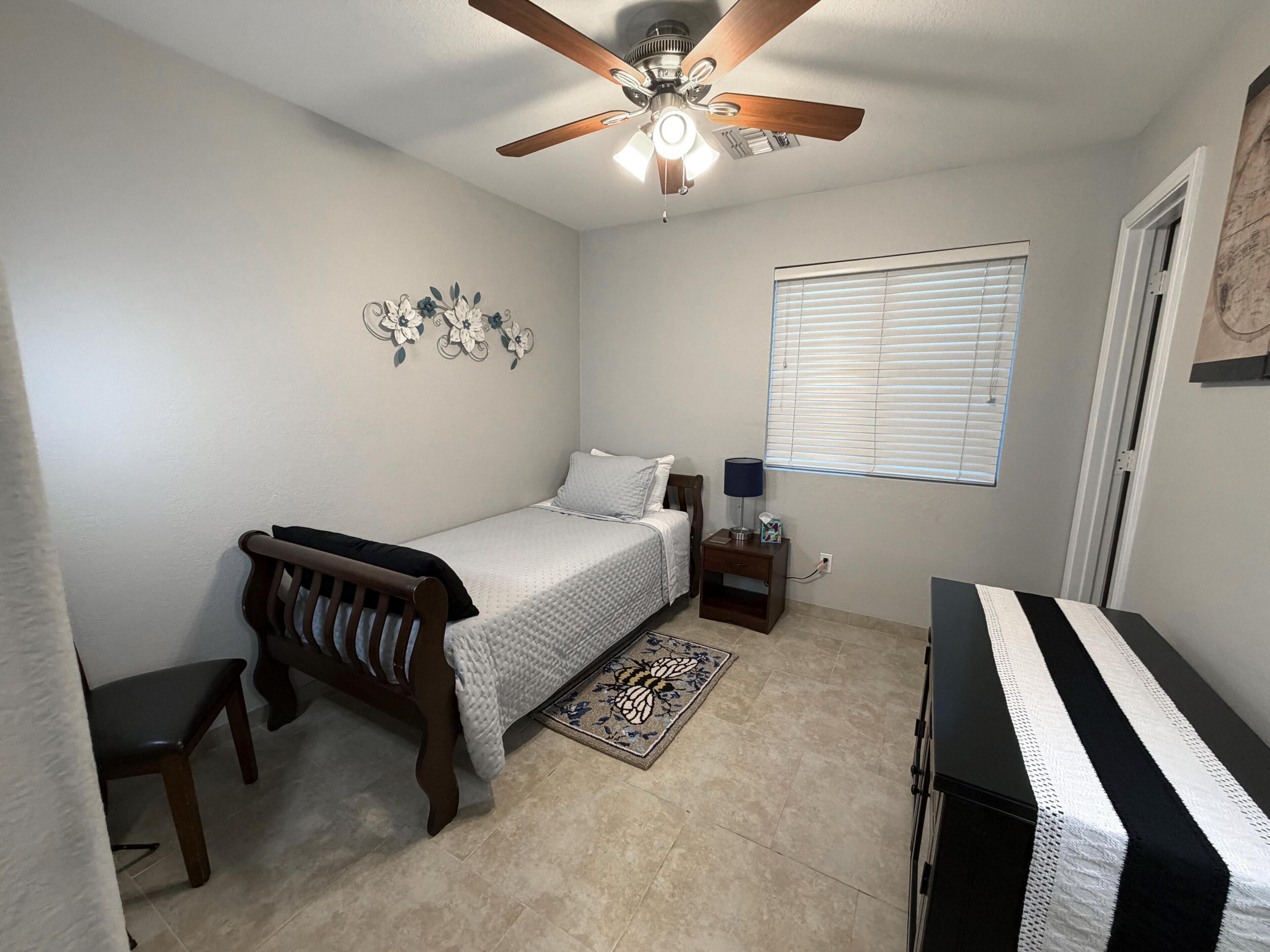 A bedroom with a bed, a nightstand, a chair, a ceiling fan, a window with blinds, and a door.