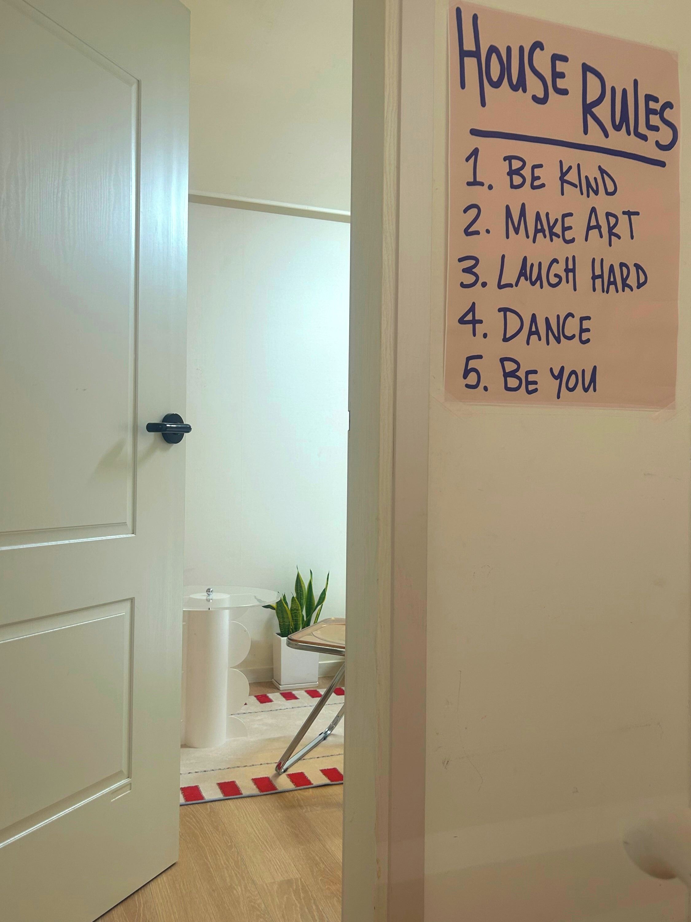 A hallway with a door leading to a room, a hand-written list titled 'House Rules' on the wall, and a plant in a white pot.