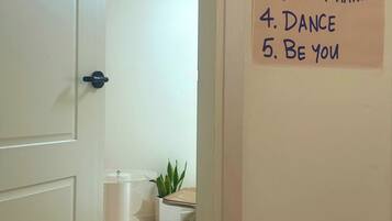 A hallway with a door leading to a room, a hand-written list titled 'House Rules' on the wall, and a plant in a white pot.