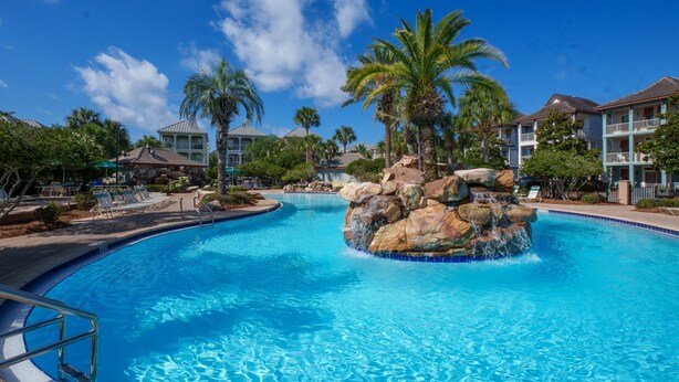 Una piscina con una roccia decorativa, palme e gli edifici di un resort.