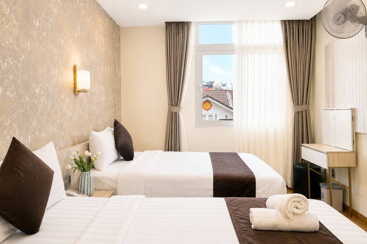 A hotel room with two beds, a bedside table, a lamp, a fan, and a window with curtains.