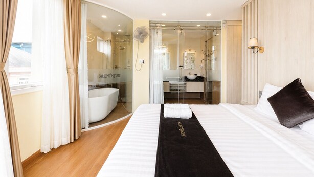 A modern hotel room with a large bed, a bathroom with a bathtub, and a view of the city. - Silentnight - Đêm Lành Hotel (Xuan Huong - Da Lat)