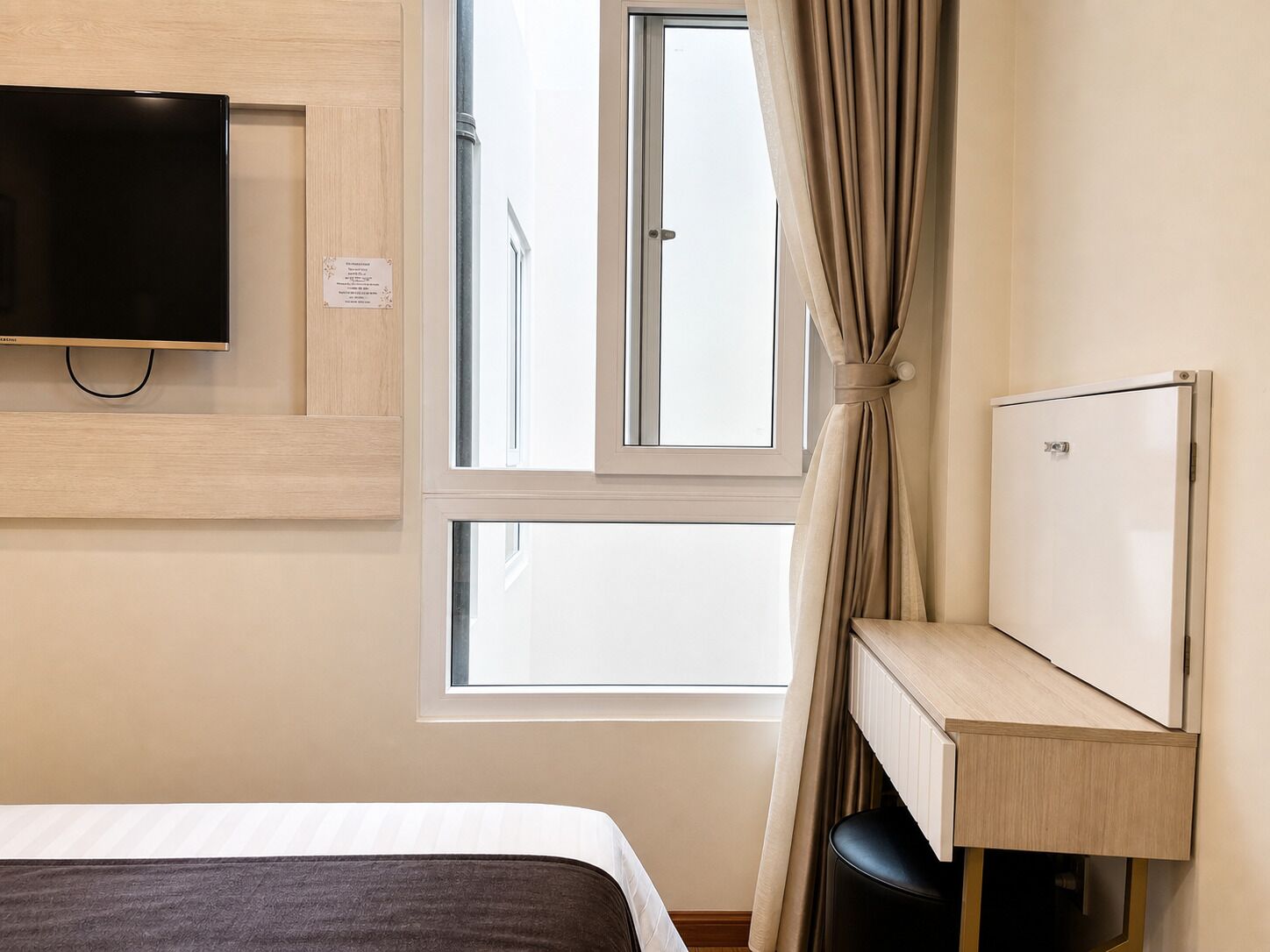 Habitación de hotel con cama, televisión montada en la pared, ventana con cortinas y una pequeña mesa de escritorio con una silla.