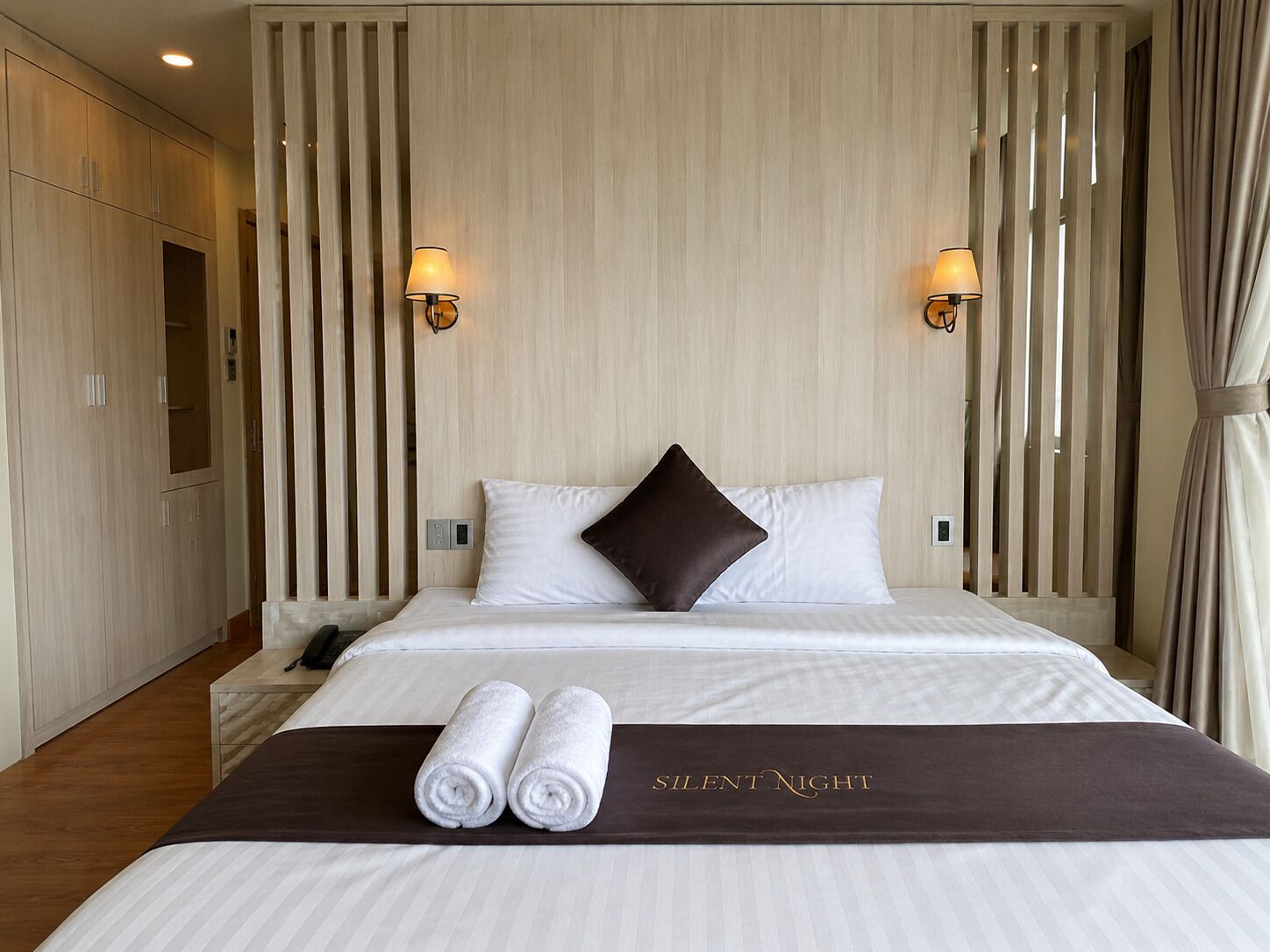 Una habitación de hotel con una cama, dos lámparas fijadas a la pared, un cabecero oscuro y un cartel en la cama que dice 'Noche Silenciosa'.