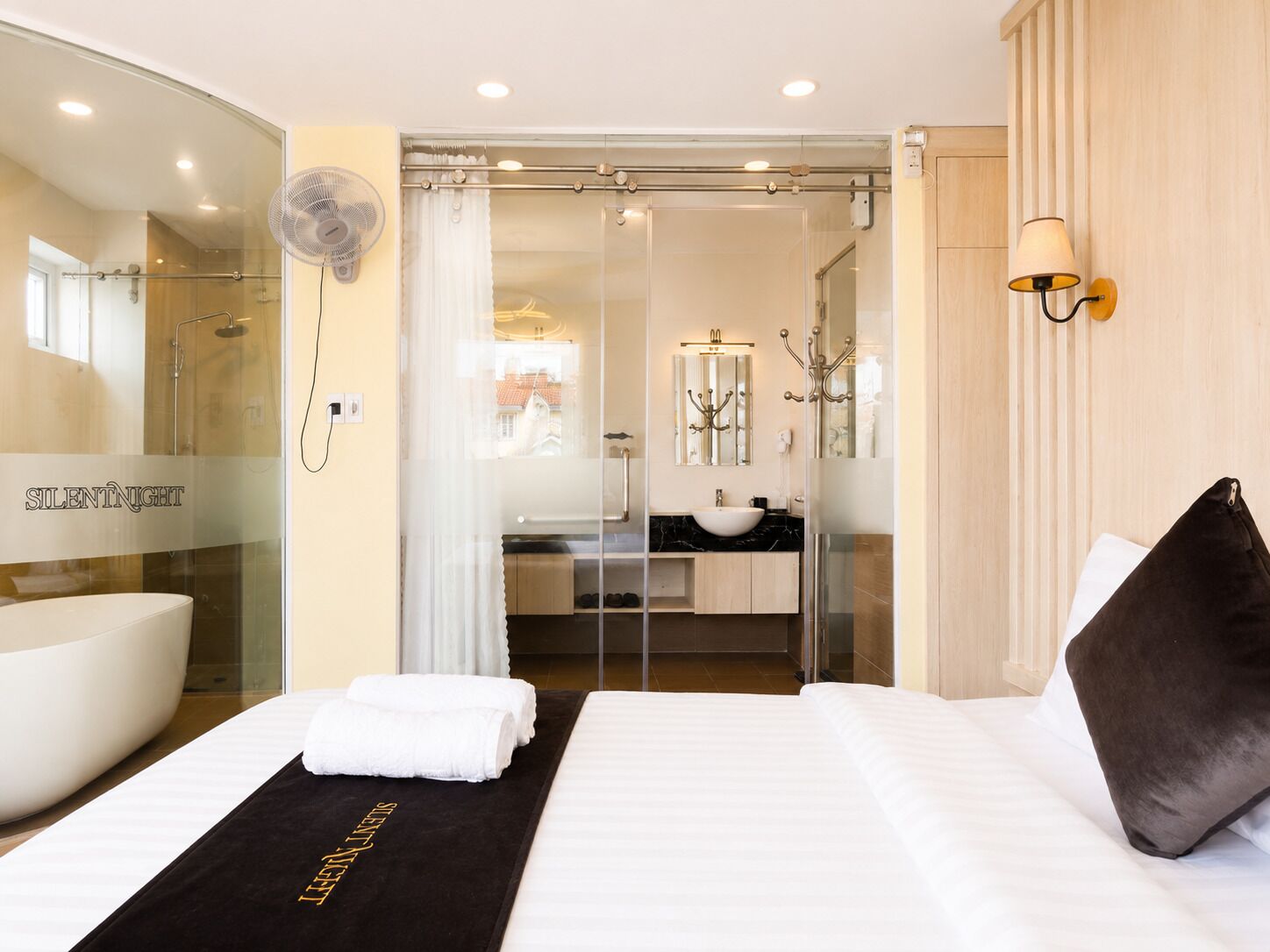 Habitación de hotel con cama, cabina de ducha de vidrio, baño con lavamanos y espejo, y ventilador de pared.