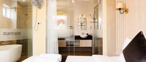 Habitación de hotel con cama, cabina de ducha de vidrio, baño con lavamanos y espejo, y ventilador de pared.