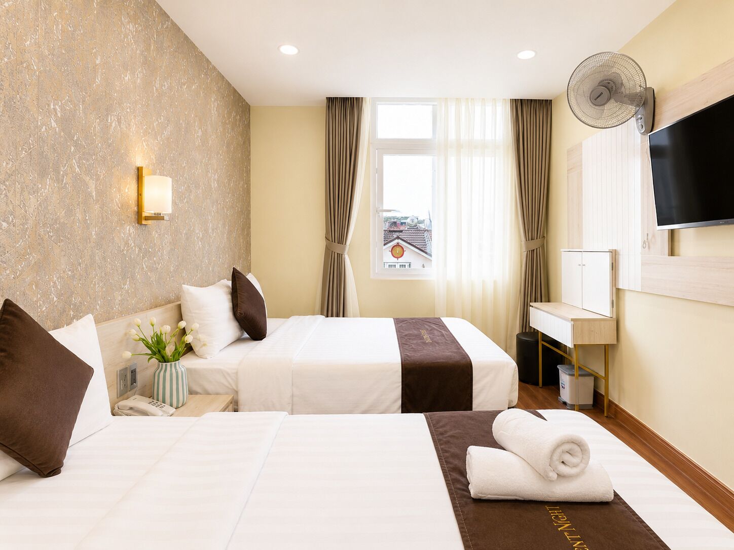 A hotel room with two beds, a TV, a fan, a window with curtains, and a view of buildings outside.