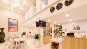 A hotel reception area with a reception desk, a framed picture on the wall, and a dining area with tables and chairs. - Silentnight - Đêm Lành Hotel (Xuan Huong - Da Lat)