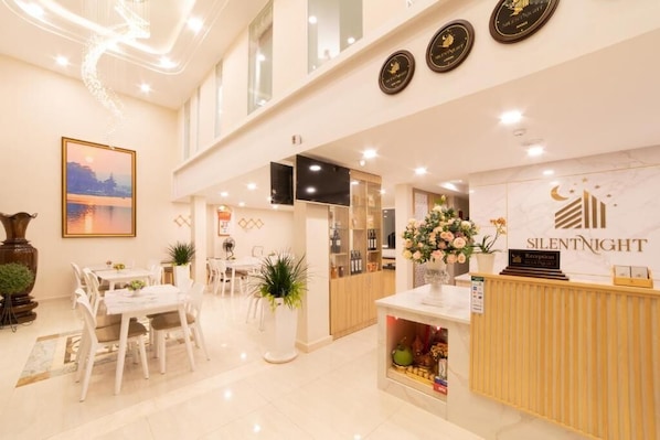 Ein Hotel-Rezeptionsbereich mit Empfangstresen, einem gerahmten Bild an der Wand und einem Essbereich mit Tischen und Stühlen.