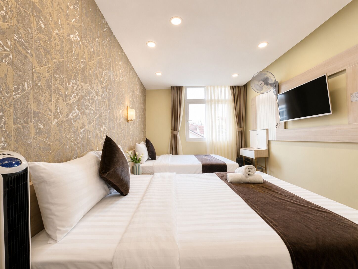 A hotel room with two beds, a TV, a fan, and a window with curtains.
