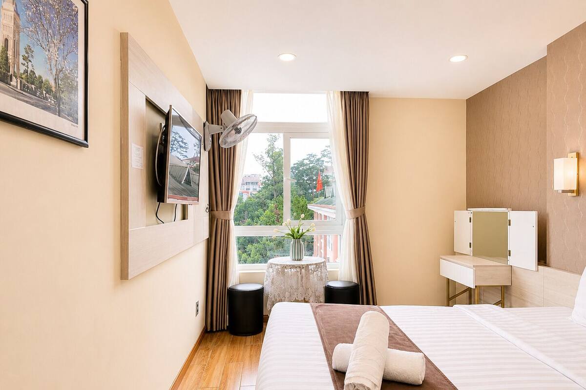 A hotel room with a bed, a TV, a window with curtains, a small table with a vase, and a mirror.