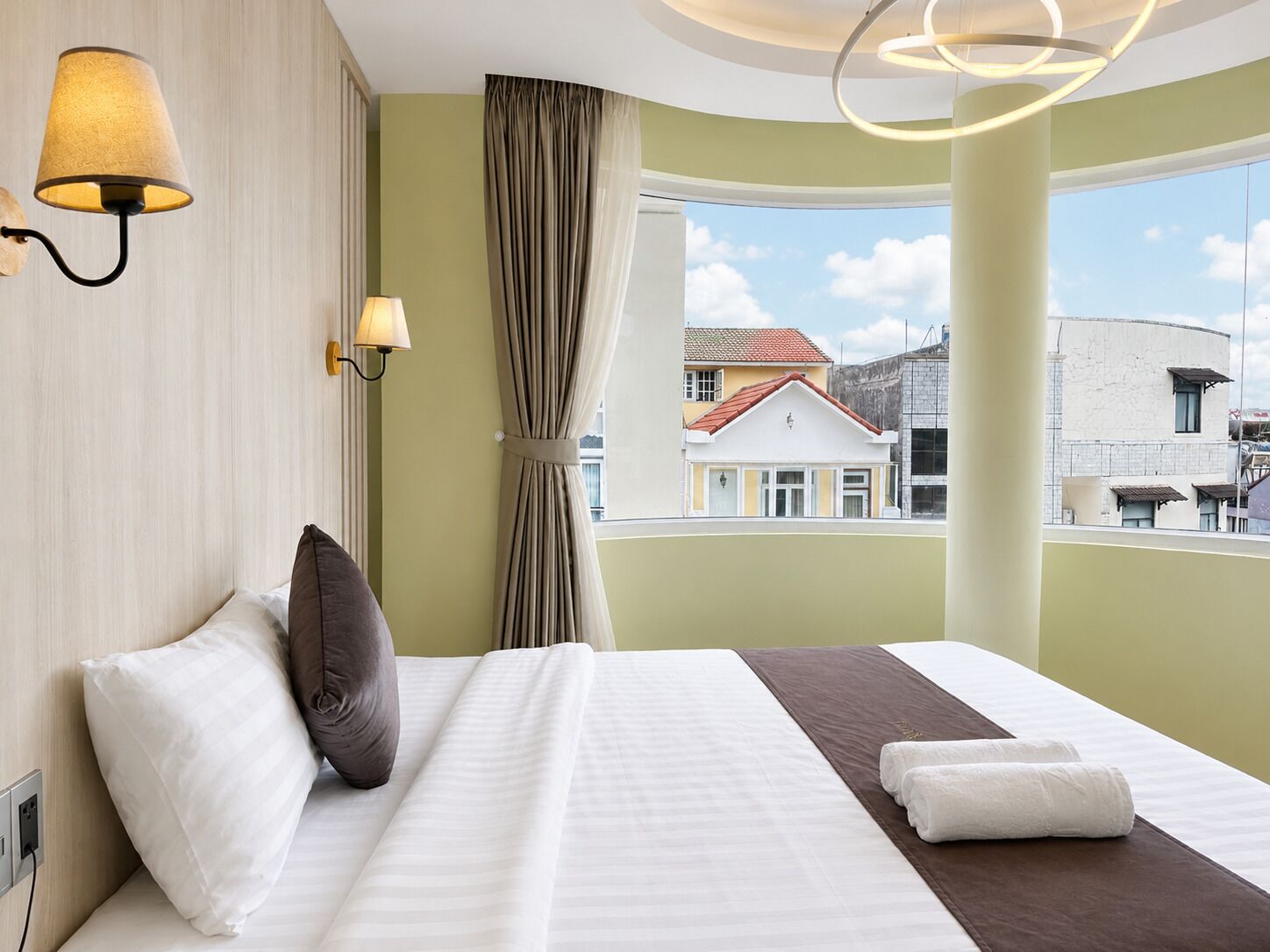 A hotel room with a large bed, two wall-mounted lamps, a window with a view of buildings, and a curtain.