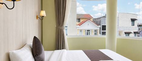 A hotel room with a large bed, two wall-mounted lamps, a window with a view of buildings, and a curtain.