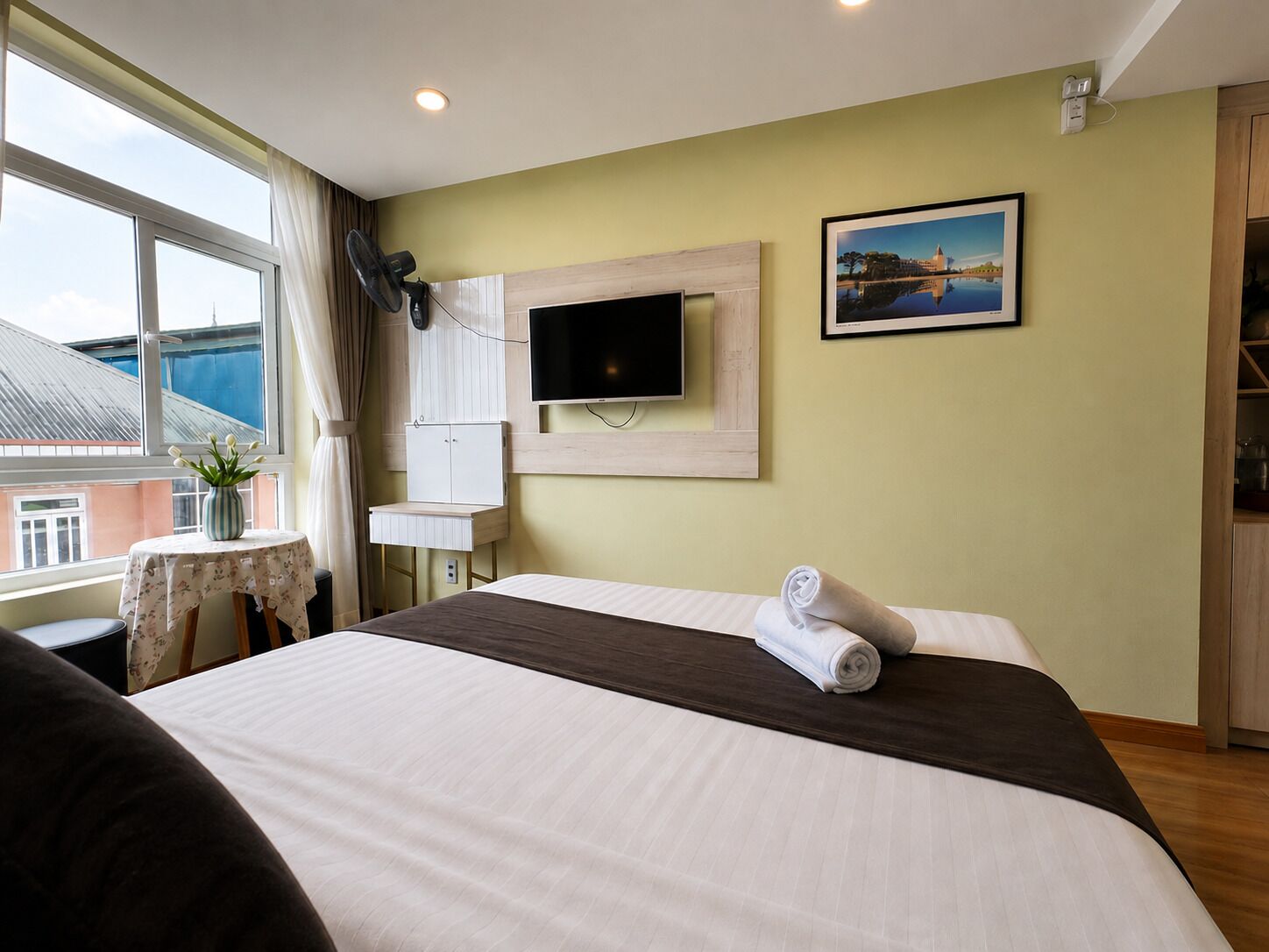 A hotel room with a bed, a TV, a painting, a window, and a small table with a vase.