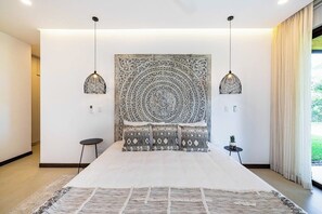 Une chambre moderne avec un grand lit, deux lampes suspendues, une tête de lit décorative et une vue sur la verdure à l’extérieur.